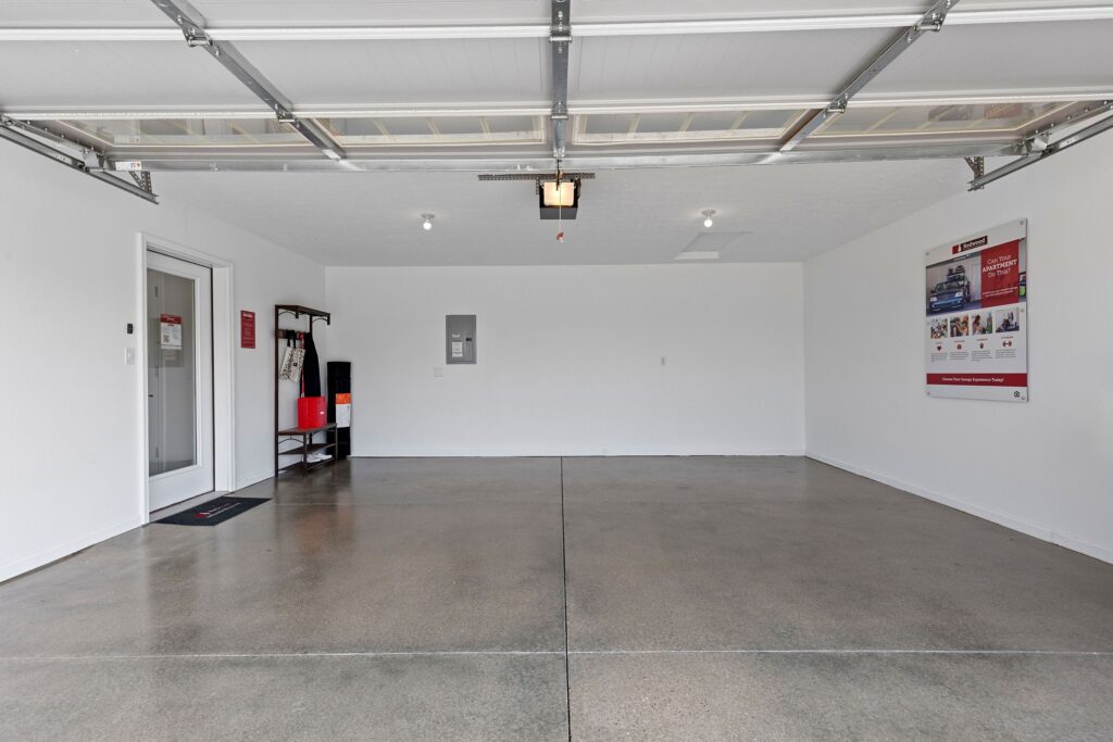A clean, empty two-car garage with white walls, a polished concrete floor, and a storage shelf on the left side. Posters are mounted on the right wall.