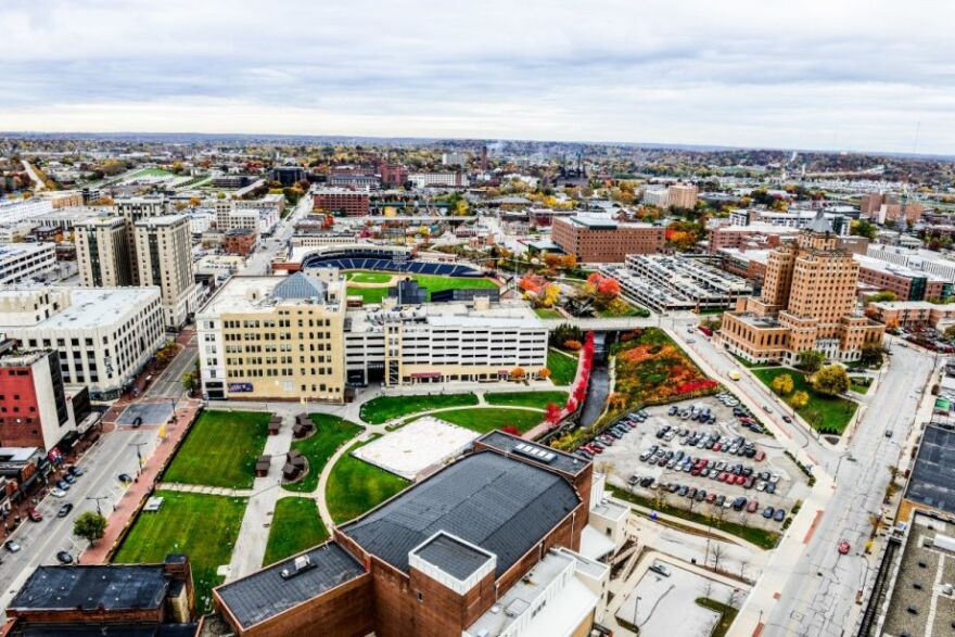 Aerial view of a cityscape featuring a stadium, parking lot, and various buildings surrounded by roads and greenery under a cloudy sky.