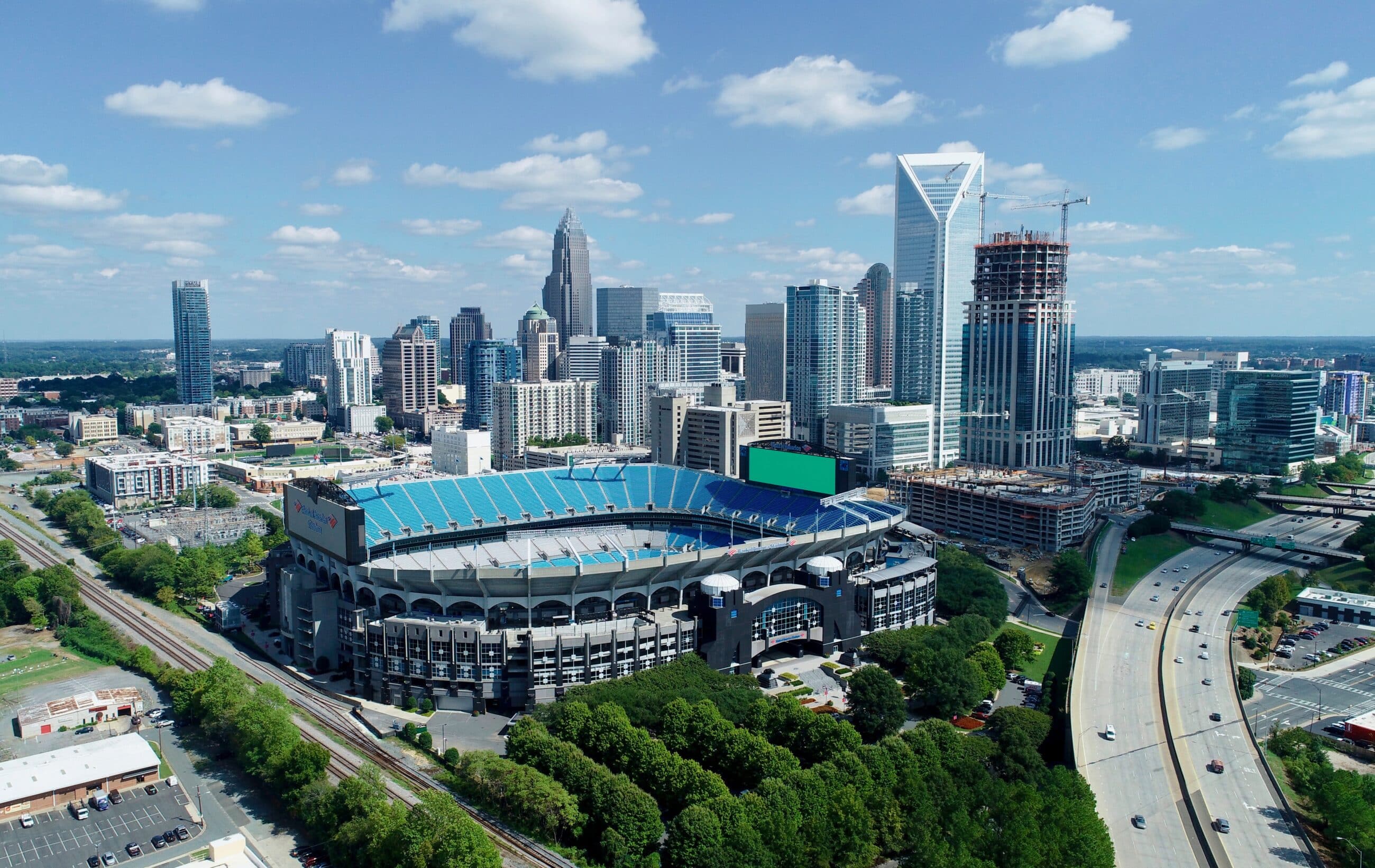 Aerial view of a stadium in a cityscape with tall buildings, roads, and greenery under a partly cloudy sky.