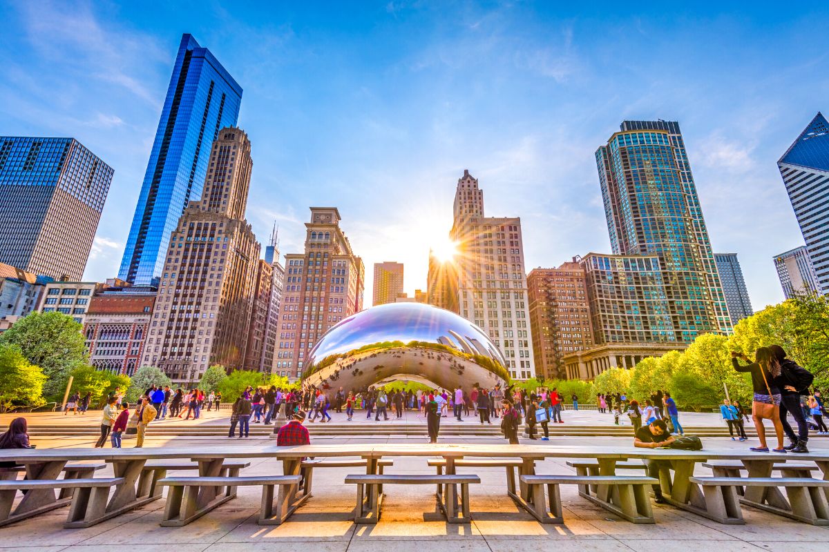 People gather around the reflective metallic sculpture, Cloud Gate, with city skyscrapers and trees in the background under a sunny, blue sky.