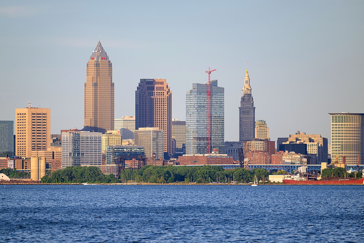 Skyline of a city with a mix of modern and historic skyscrapers under a clear sky, viewed from across a body of water, with trees in the foreground.