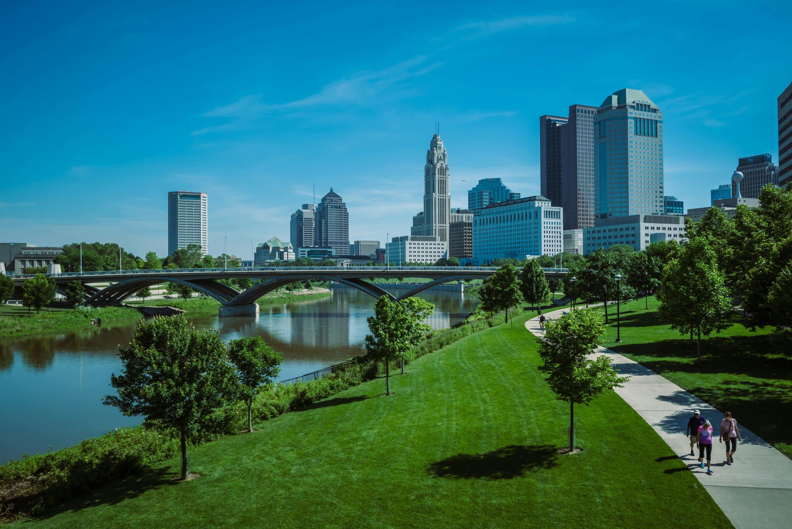 Cityscape view with skyscrapers, a river, and a bridge. A path runs through a green park with people walking. Clear blue sky overhead.