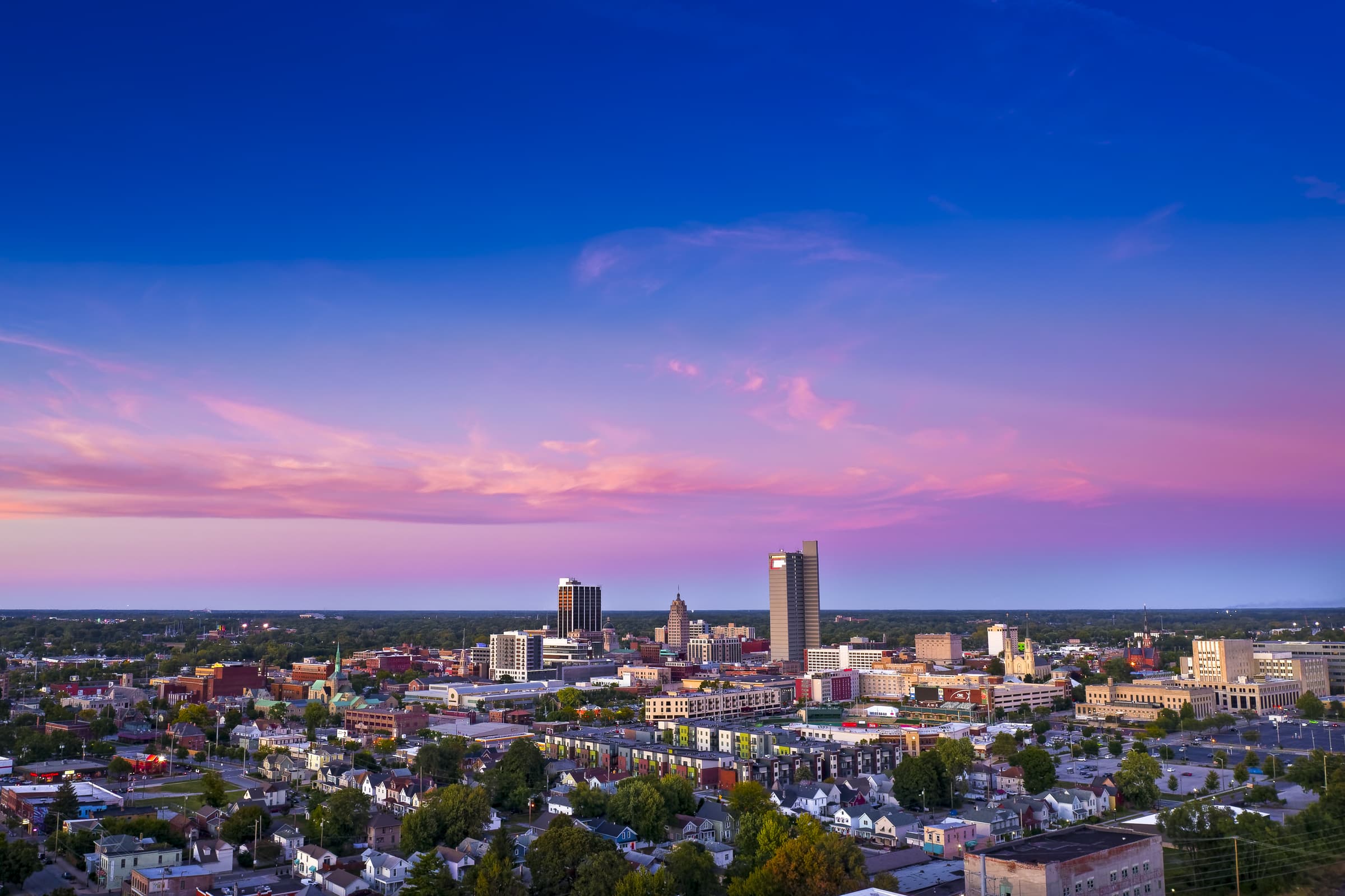 Aerial view of a cityscape at dusk, featuring a mix of tall buildings, houses, and trees under a pink and blue sky.