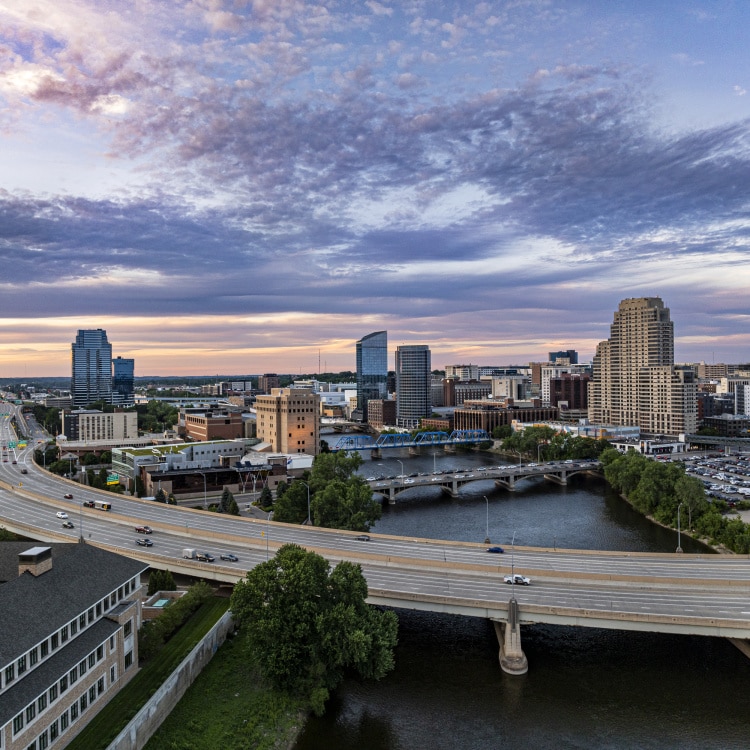 Aerial view of a city skyline with tall buildings, a river, and a bridge. The sky is partly cloudy during sunset.