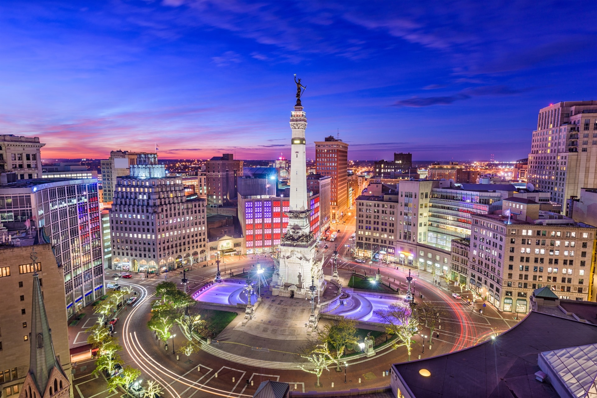 Aerial view of Monument Circle in downtown Indianapolis at dusk, with the Soldiers and Sailors Monument surrounded by city buildings and light trails from traffic.