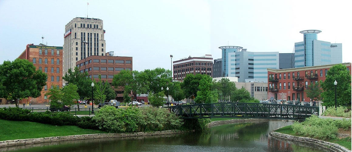 A small river with a bridge in the foreground, surrounded by green trees and grass. Urban buildings of varying heights stand in the background under a cloudy sky.