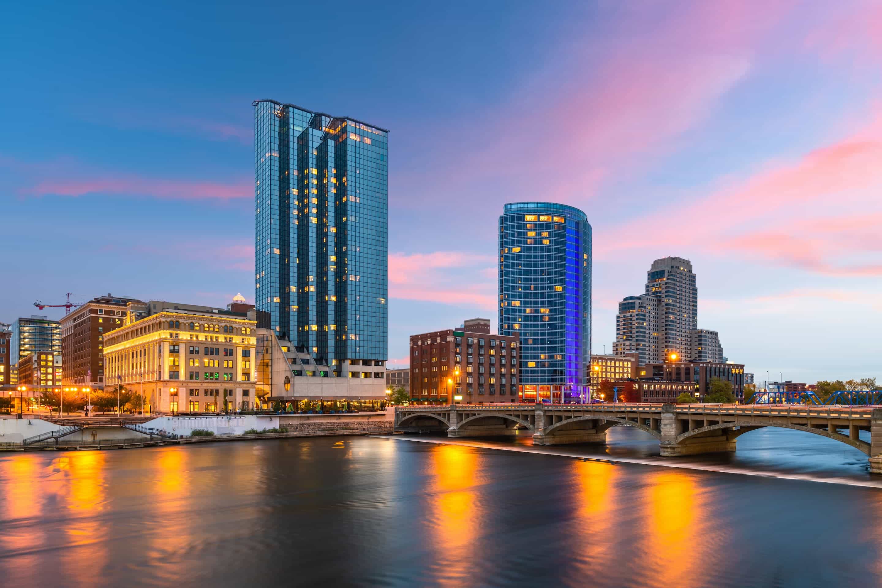 City skyline with modern and historic buildings, a river, and a bridge at sunset, with reflections of lights on the water.