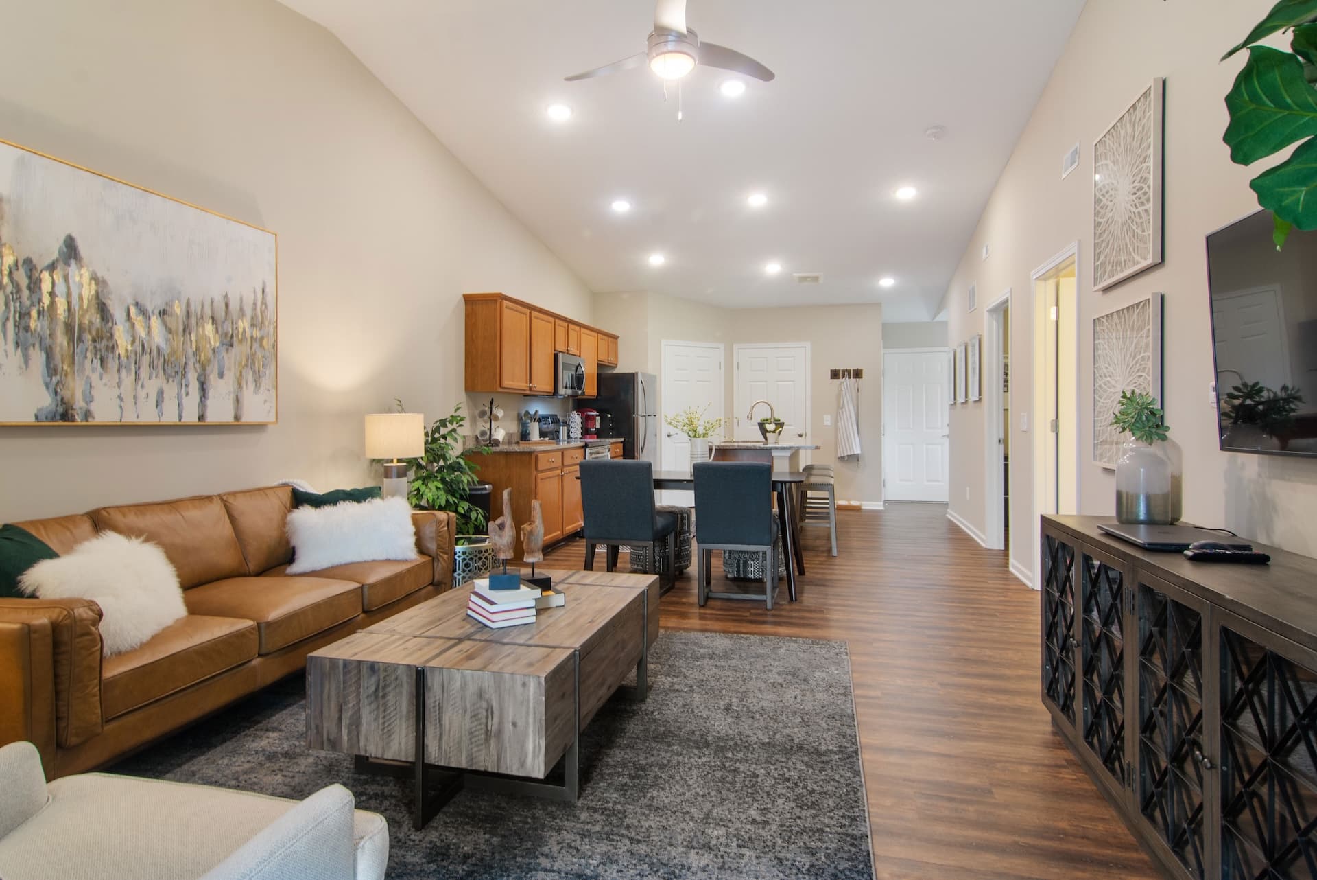 Open-concept living room and kitchen with brown leather sofa, wooden dining table, modern decor, ceiling fan, and hardwood floors.