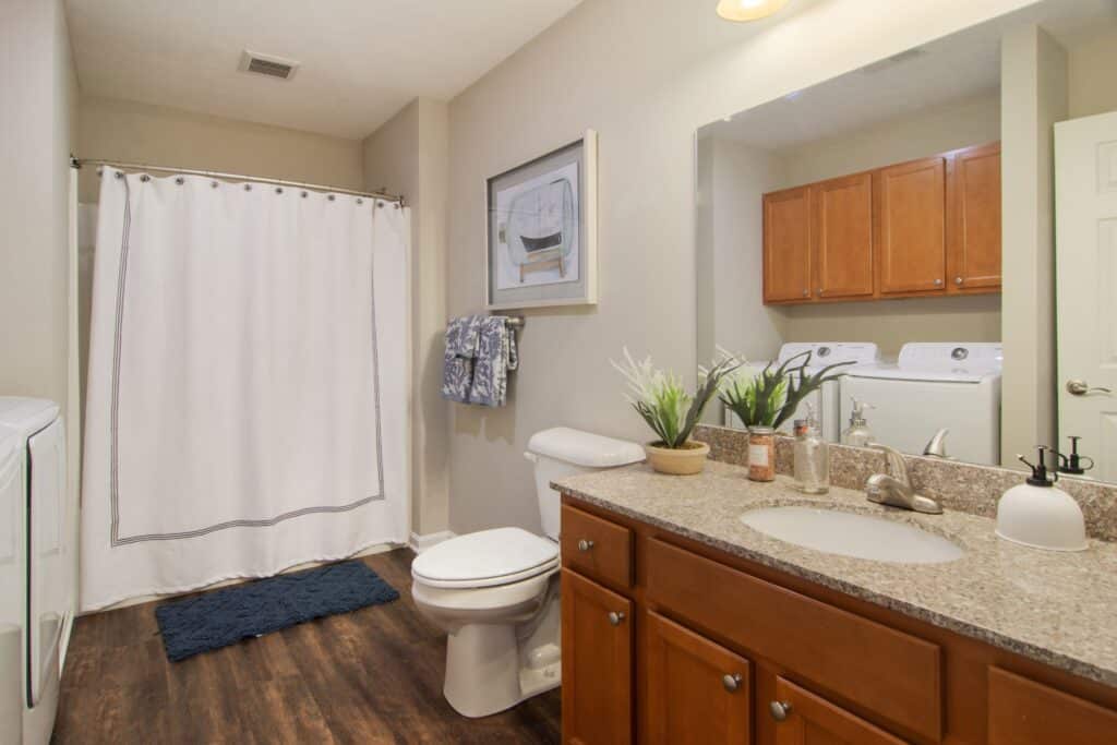 Bathroom with wooden cabinets, granite countertop, toilet, shower with curtain, washer and dryer, and decorative items on the counter.