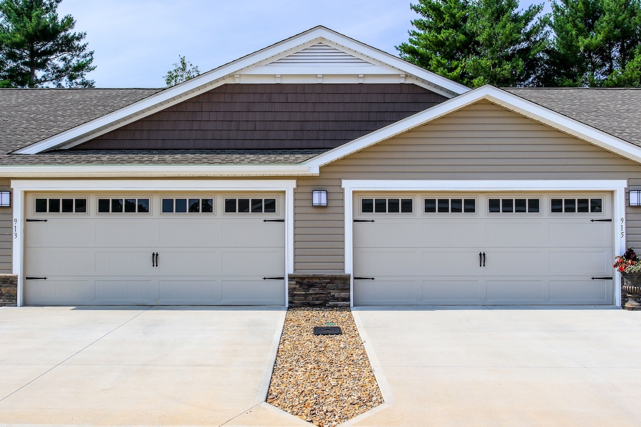 A duplex building with two white garage doors, beige siding, and a shared stone-lined driveway divider, surrounded by trees.