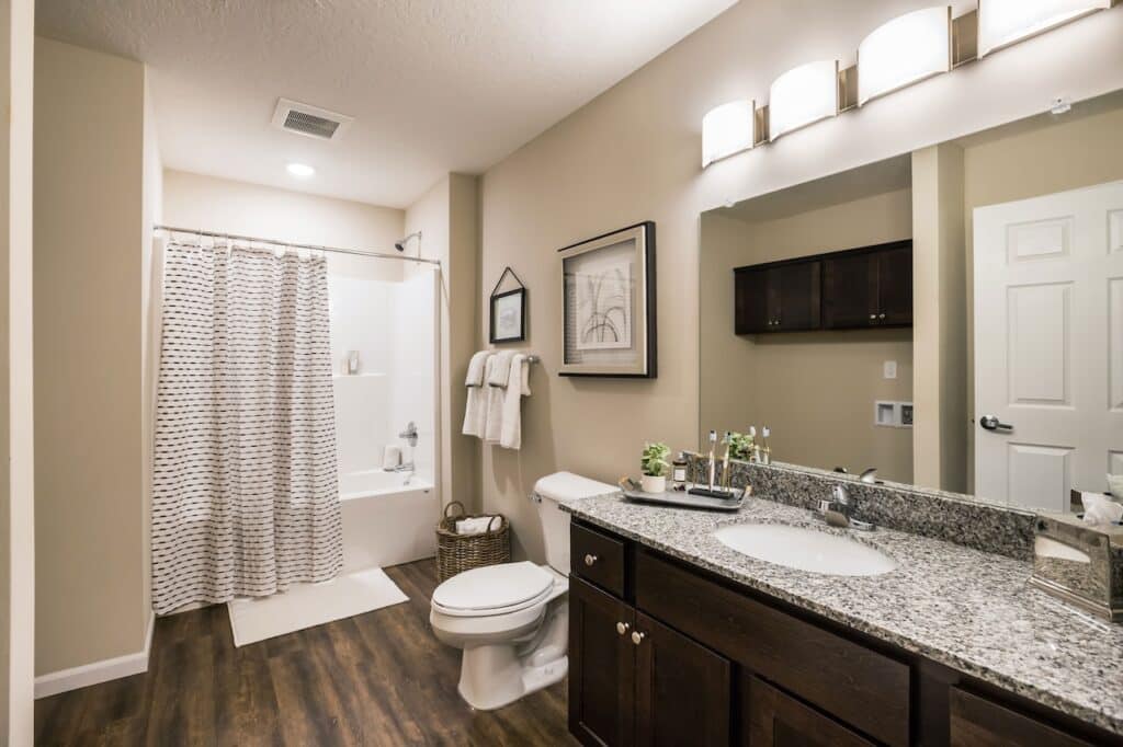 Modern bathroom with granite countertop, dark wood cabinets, a toilet, a bathtub with a patterned shower curtain, wall art, and wood-look flooring under bright lighting.