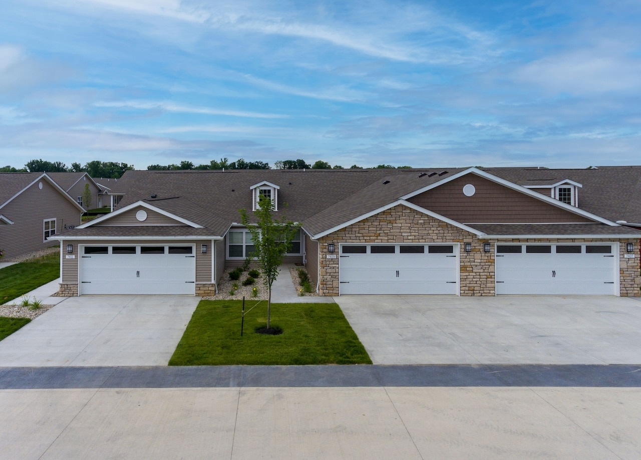 A row of modern, single-story duplex homes with attached two-car garages and concrete driveways under a partly cloudy sky.