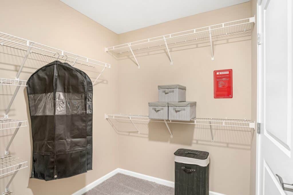 Organized closet with wire shelves, a black garment bag, three gray storage boxes, a black hamper, and a red sign on a beige wall.