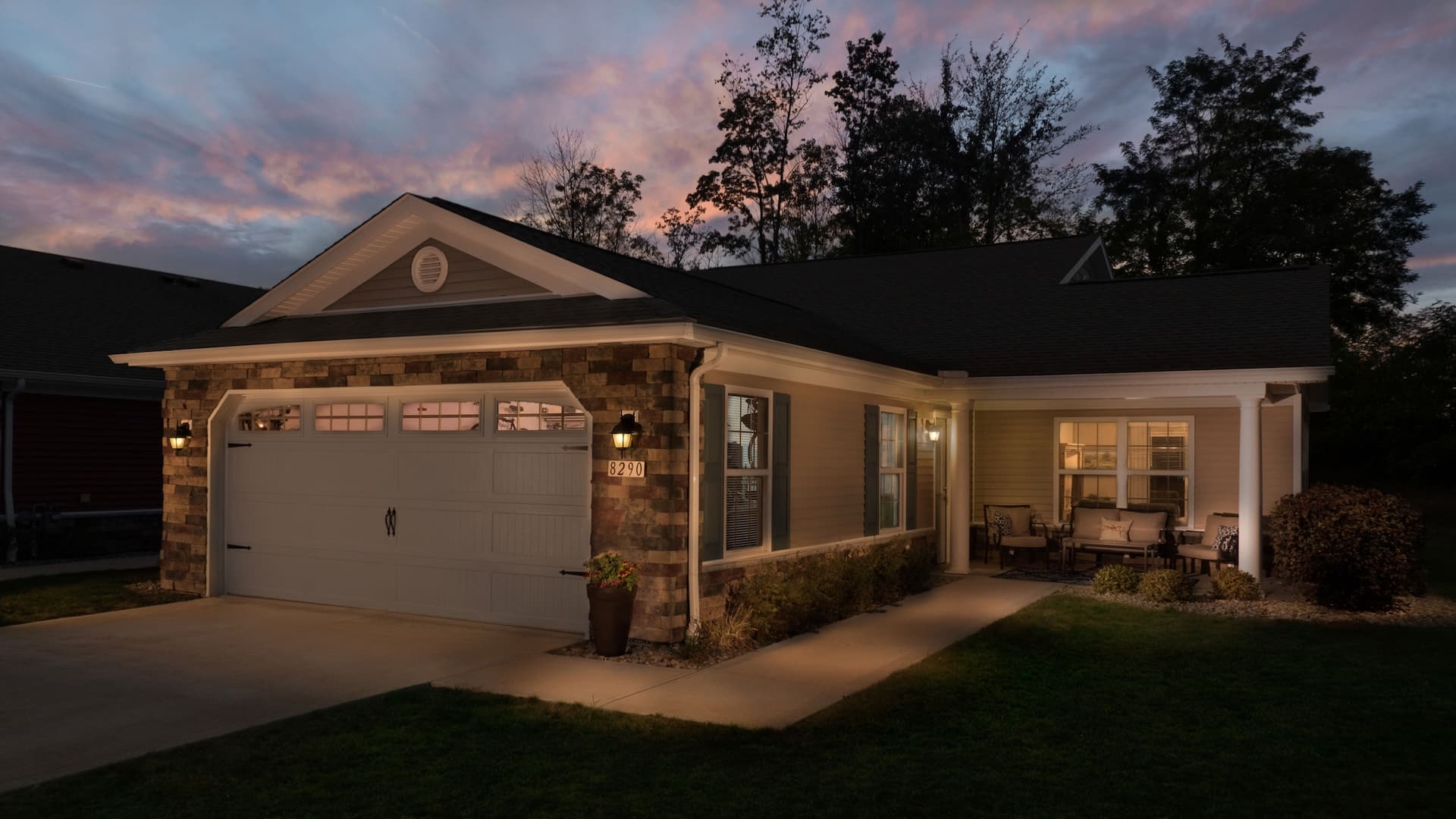 Single-story house with stone and siding exterior, lit porch, two-car garage, outdoor seating area, and landscaped front yard at dusk.