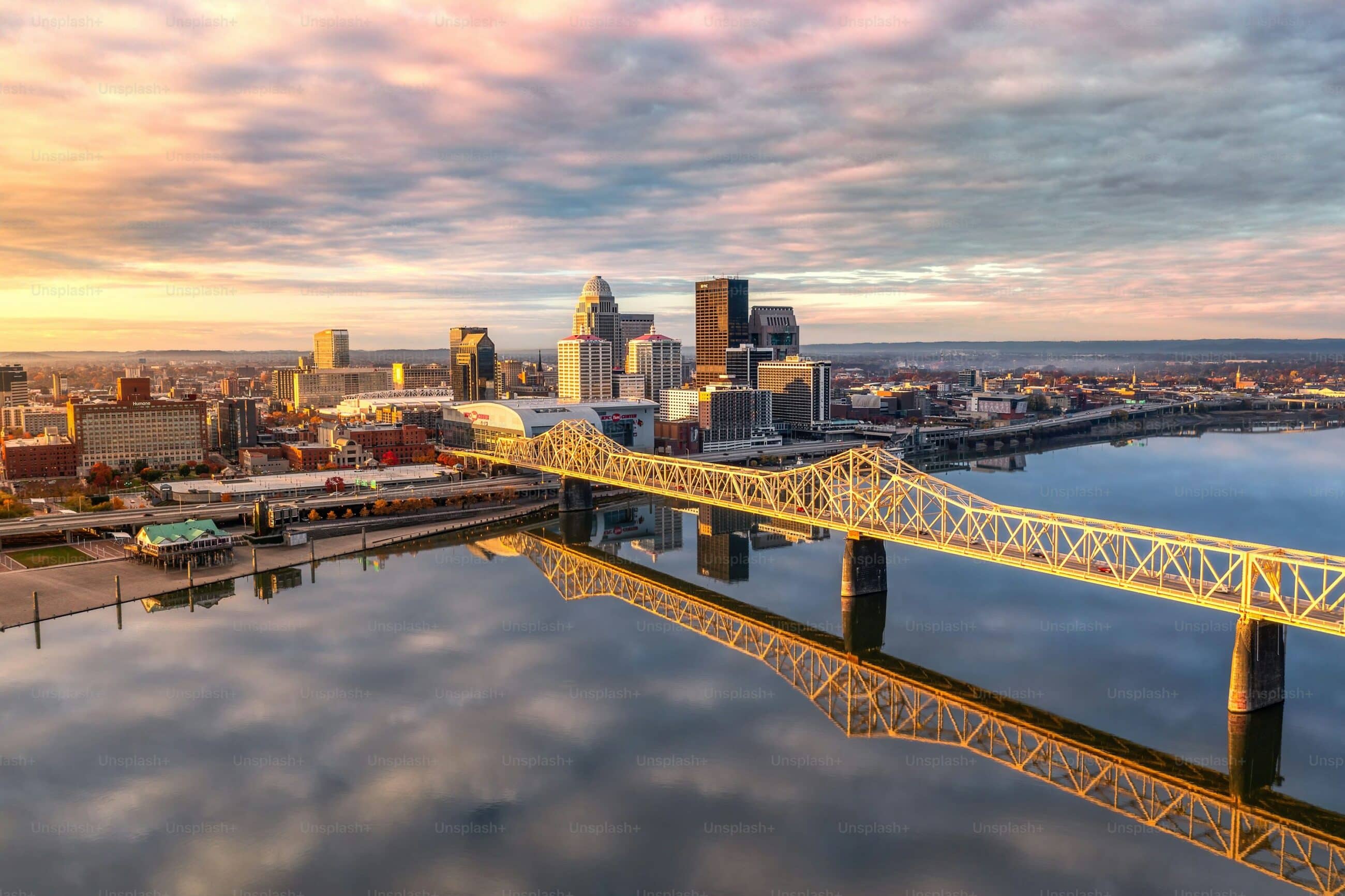 Cityscape view of a downtown area with a prominent bridge spanning a wide river. The skyline includes modern high-rise buildings under a partly cloudy sky reflecting on the water.