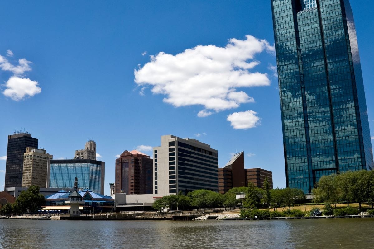 Skyline of a city featuring modern high-rise buildings, a blue sky with scattered clouds, and a river in the foreground.
