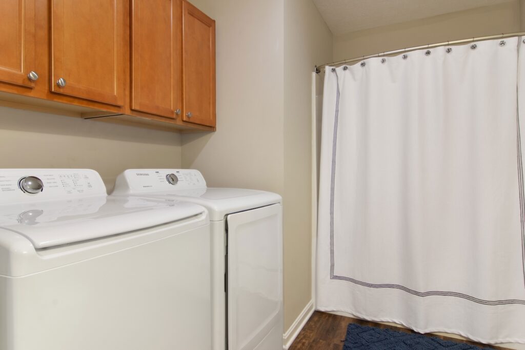 A laundry room with a washing machine and dryer side by side, wooden cabinets above, and a shower with a white curtain in the background.