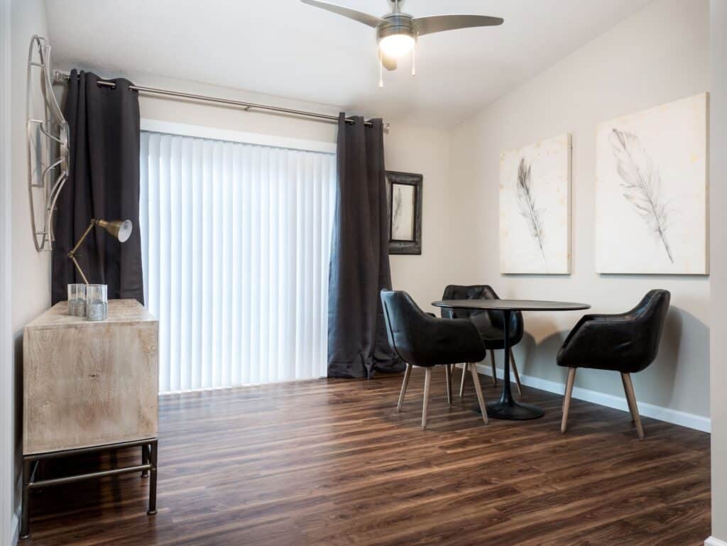A modern dining area with a round table, four black chairs, wooden sideboard, large windows with blinds, dark curtains, and two feather artworks on the wall.