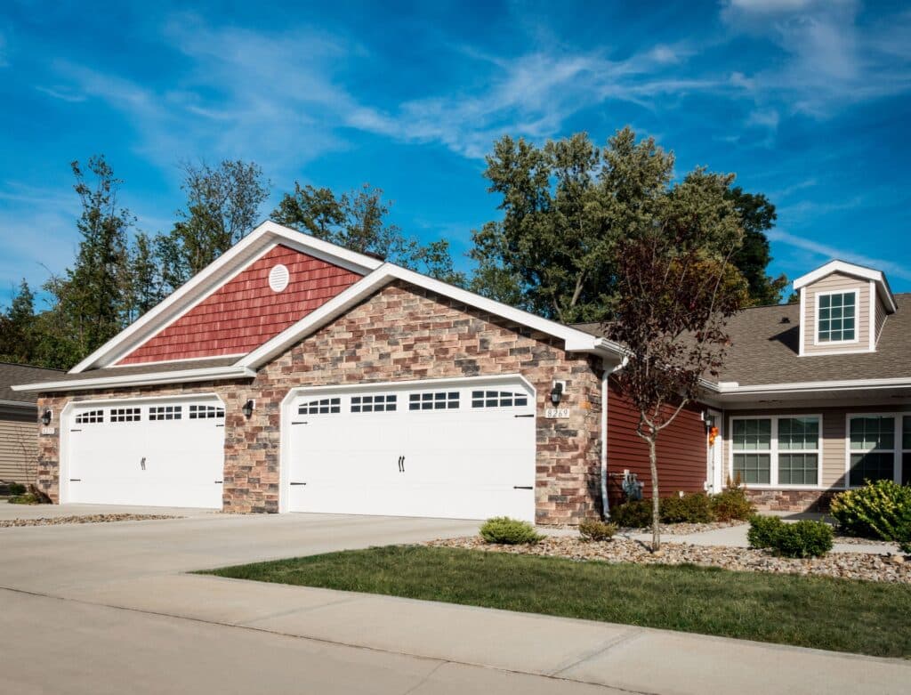 A suburban duplex with brick and red siding exterior, featuring two large white garage doors, a manicured lawn, and a clear blue sky.