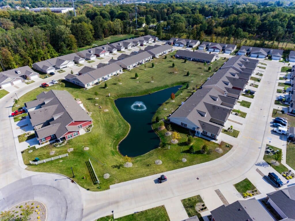 Aerial view of a suburban residential complex with rows of single-story homes surrounding a small pond with a water fountain, green lawns, and paved roads.