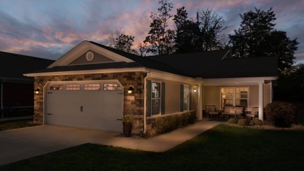 Single-story house with illuminated exterior lights, stone and siding facade, double garage, small front porch, and manicured lawn at dusk.