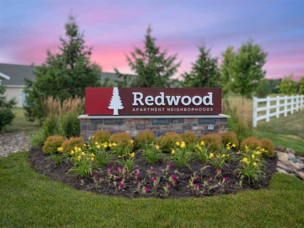 A sign reading "Redwood Apartment Neighborhoods" stands in front of landscaped flowers and grass, with trees and a white fence in the background at sunset.