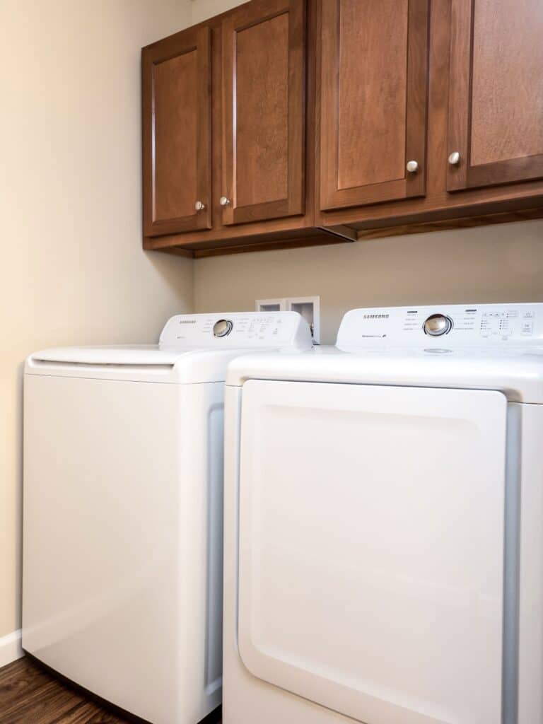 A laundry room with a white washer and dryer side by side beneath wooden cabinets mounted on a beige wall.
