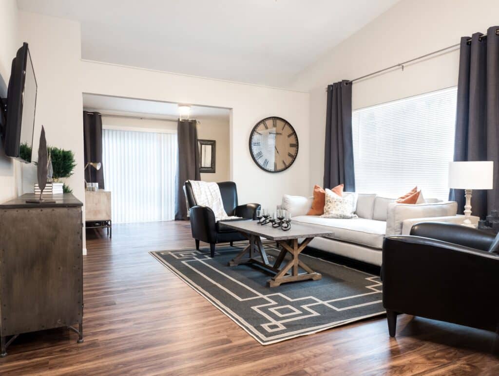 Modern living room with wood flooring, a gray sofa, two black armchairs, a coffee table, large wall clock, and floor-to-ceiling windows with dark curtains.