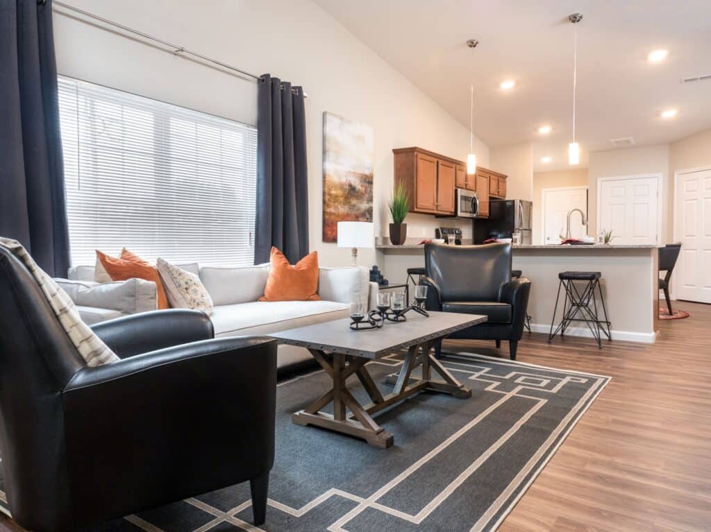 Modern living room with a gray sofa, black armchairs, coffee table, rug, and open kitchen with wooden cabinets and pendant lights in the background.