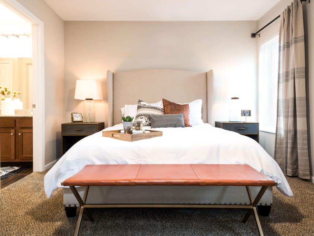 A modern bedroom with a beige upholstered bed, white bedding, assorted pillows, a tray, two dark nightstands with lamps, and a brown bench at the foot of the bed.