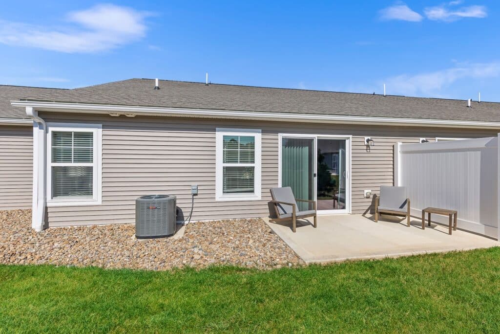 Single-story house with beige siding, sliding glass door, small concrete patio, two outdoor chairs, central AC unit, and a grassy yard under a blue sky.