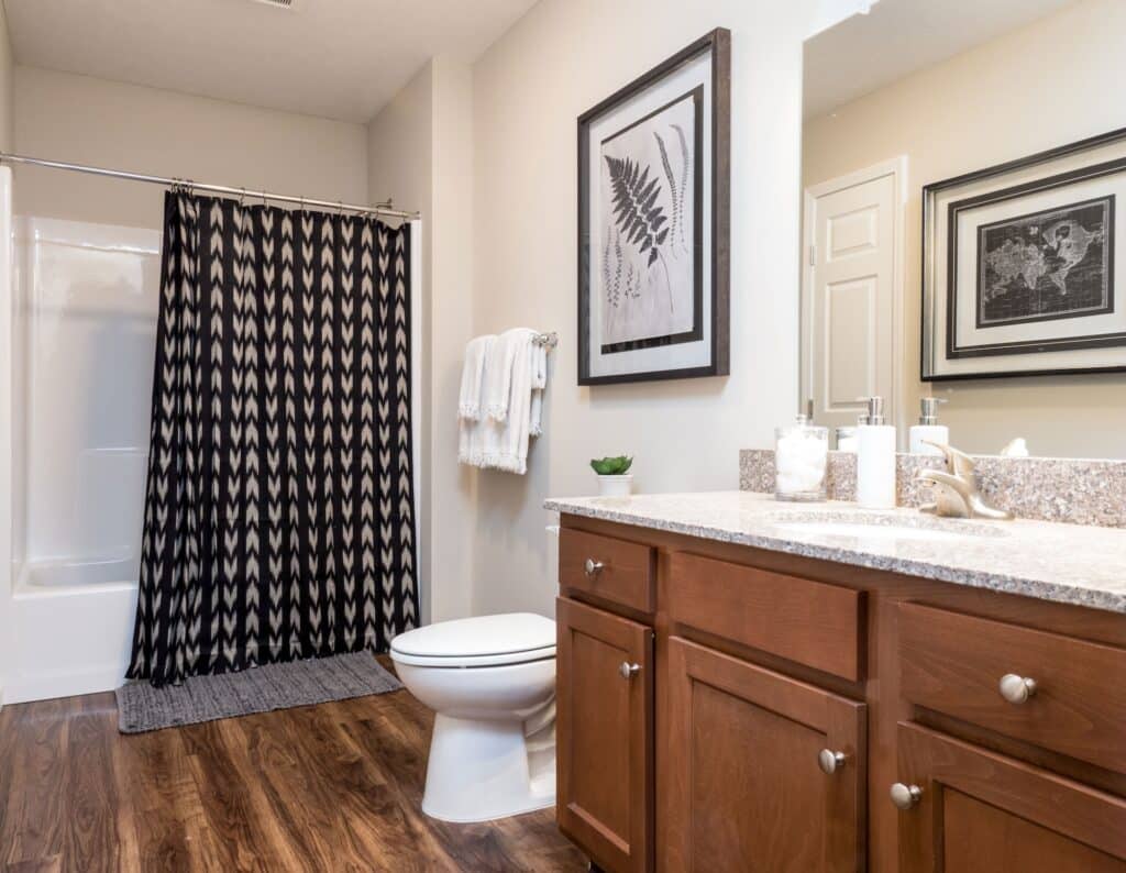 Modern bathroom with wooden cabinets, granite countertop, a toilet, a shower with a patterned curtain, wall art, towels on a rack, and wood-look flooring.