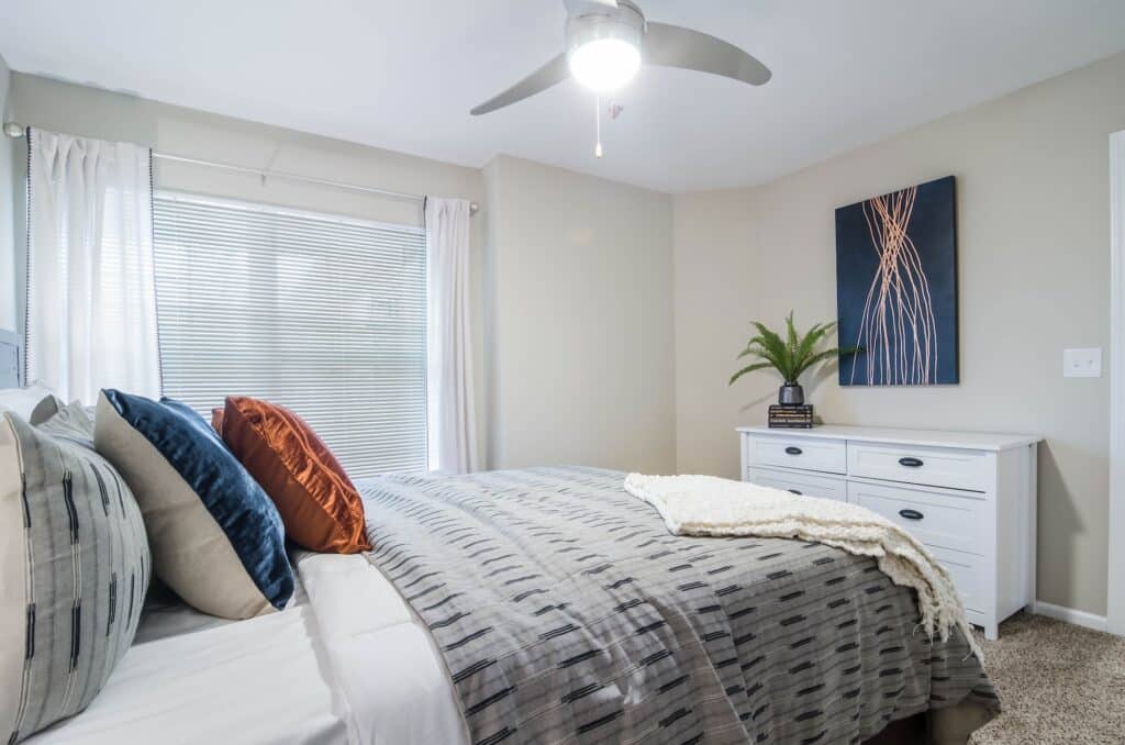 A neatly made bed with patterned bedding, colorful pillows, and a throw blanket sits beside a window. A dresser with books, a potted plant, and abstract wall art is against the wall.