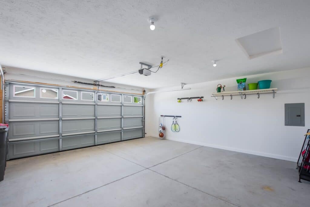 Clean, spacious two-car garage with a closed sectional door, overhead lighting, wall-mounted shelves, and organized storage items.