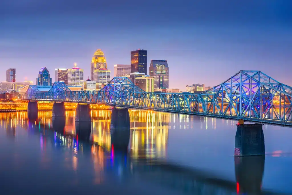 A steel truss bridge spans a calm river with city buildings illuminated in the background at dusk.