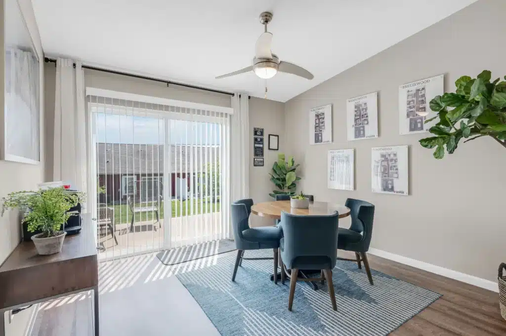 Modern dining area with round table, four blue chairs, large window with blinds, wall art, indoor plants, and a ceiling fan, overlooking a backyard through glass doors.