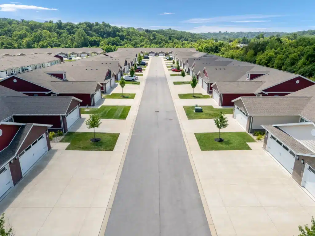 A wide street runs between rows of identical red-roofed suburban houses with garages, lawns, and a backdrop of green trees under a blue sky.