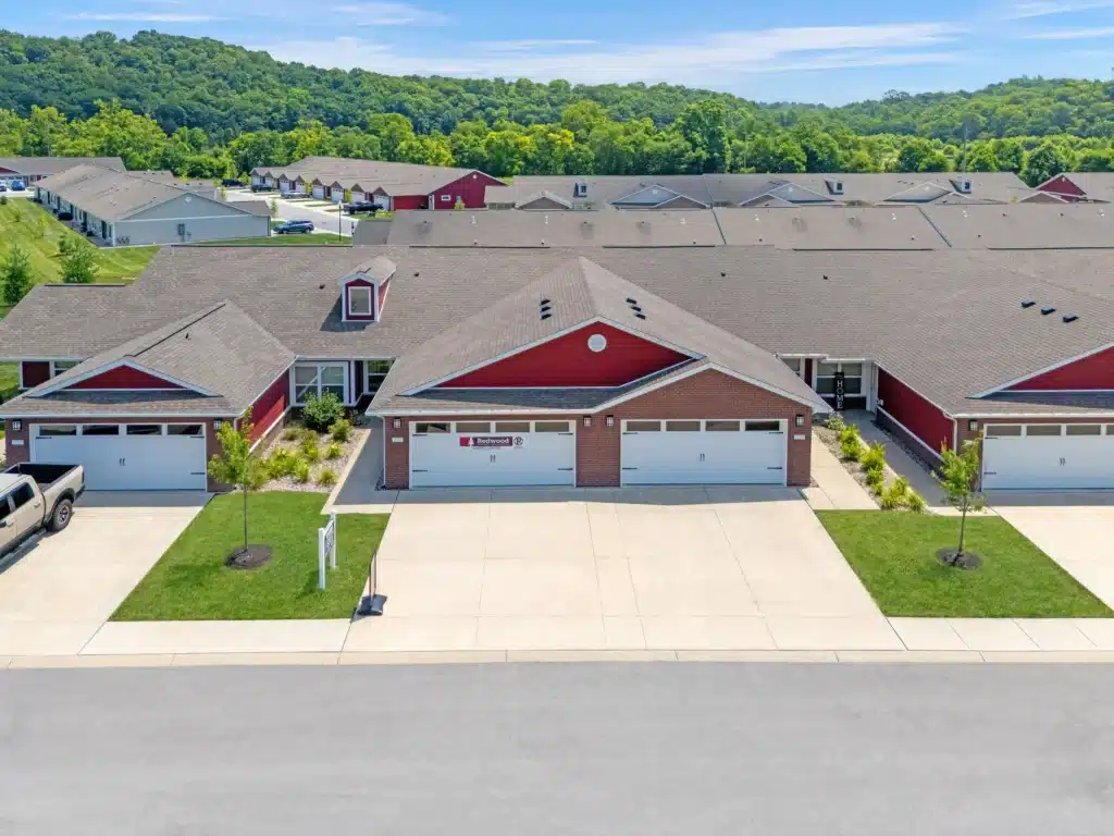 Aerial view of a row of red brick duplex homes with double garages, neat lawns, and a driveway, set against a backdrop of trees and hills.