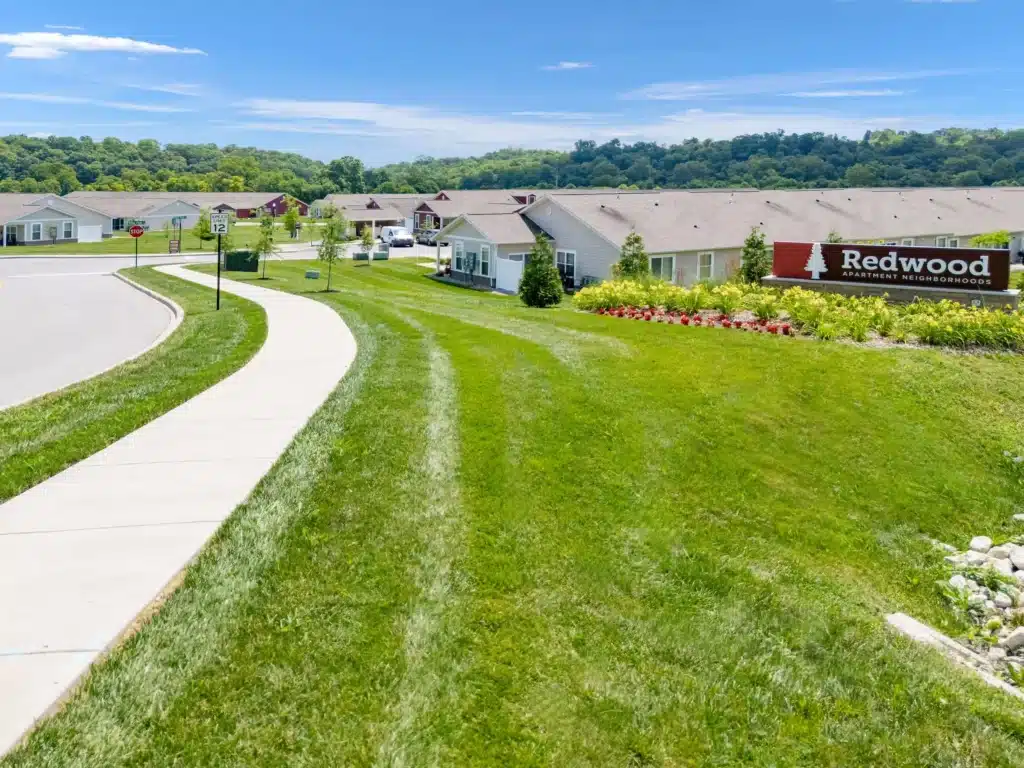 A paved sidewalk runs alongside a grassy lawn and single-story homes in a residential neighborhood with a sign reading "Redwood Apartment Neighborhood.