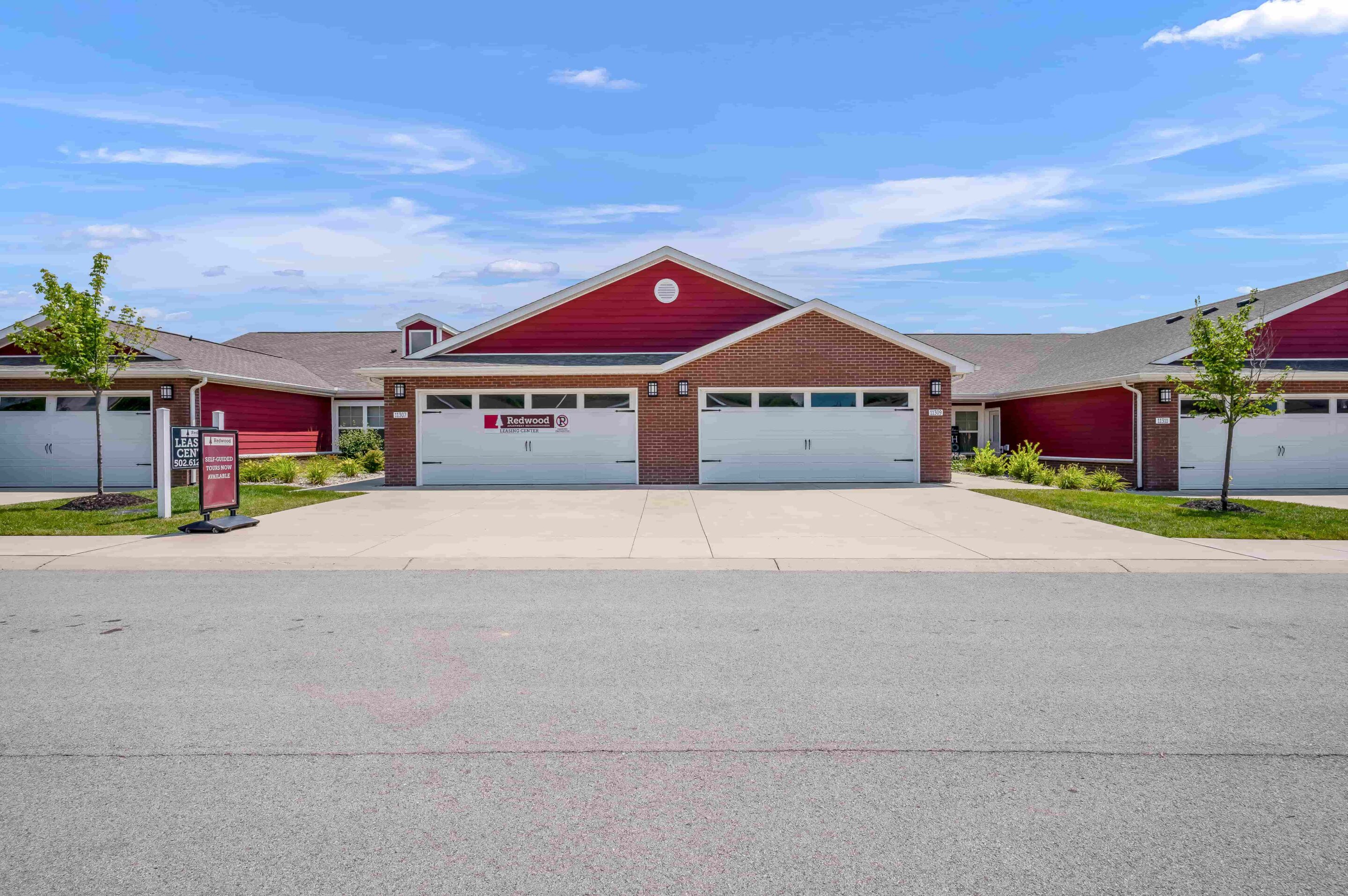 Single-story red townhouse with white trim, two garages, small front lawn, sidewalk, and a clear blue sky above.