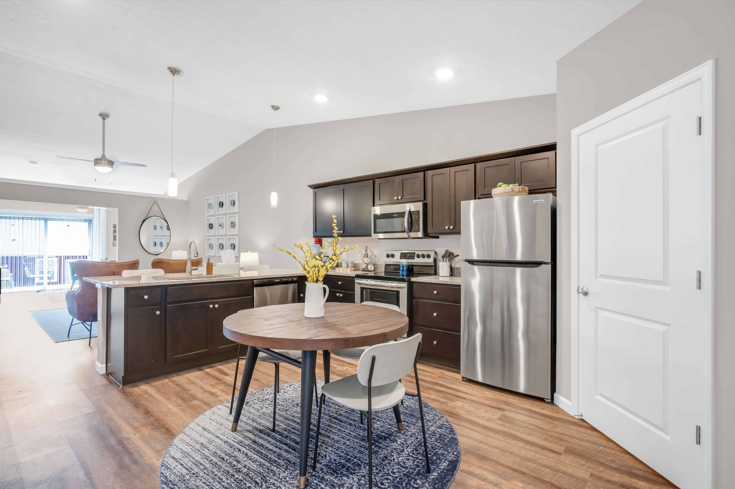 Modern kitchen and dining area with dark cabinets, stainless steel appliances, round table with a vase, wooden floors, and pendant lighting. Living area visible in the background.