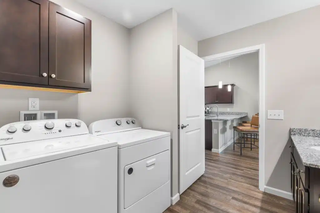 A laundry room with a white washing machine and dryer, brown upper cabinets, granite countertop, and a view into a kitchen with bar stools and wood flooring.