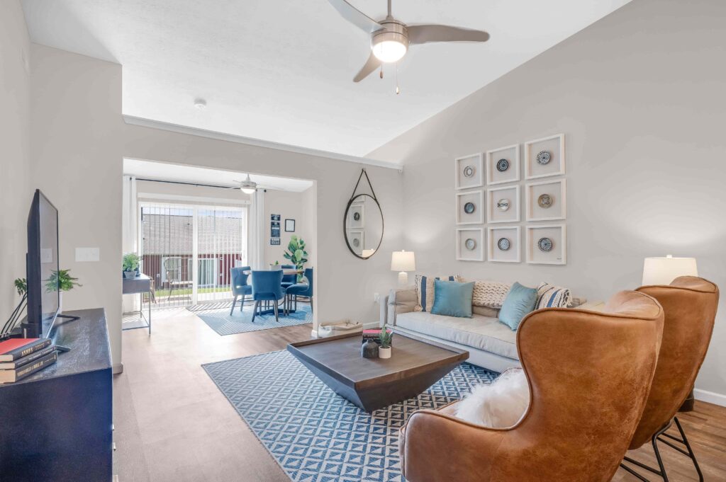 Modern living room with tan armchairs, a beige sofa, wall art, and a coffee table, opening into a dining area with a round table and large window.