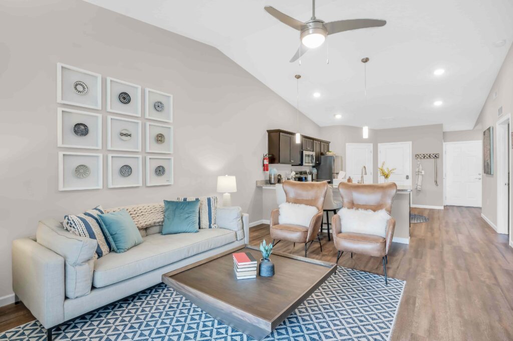 Modern living room with a beige sofa, two white chairs, a wooden coffee table, and kitchen area in the background. Neutral decor with blue accents and geometric rug.