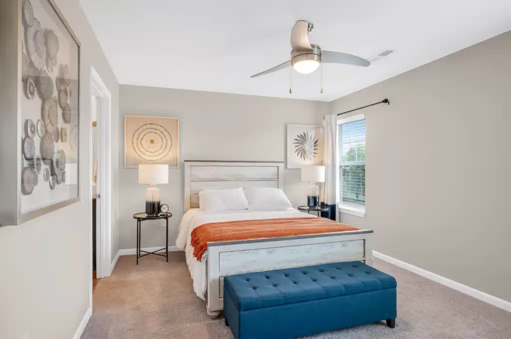 A modern bedroom with a white bed, orange throw, two bedside tables with lamps, artwork on the walls, a blue upholstered bench, and a ceiling fan.