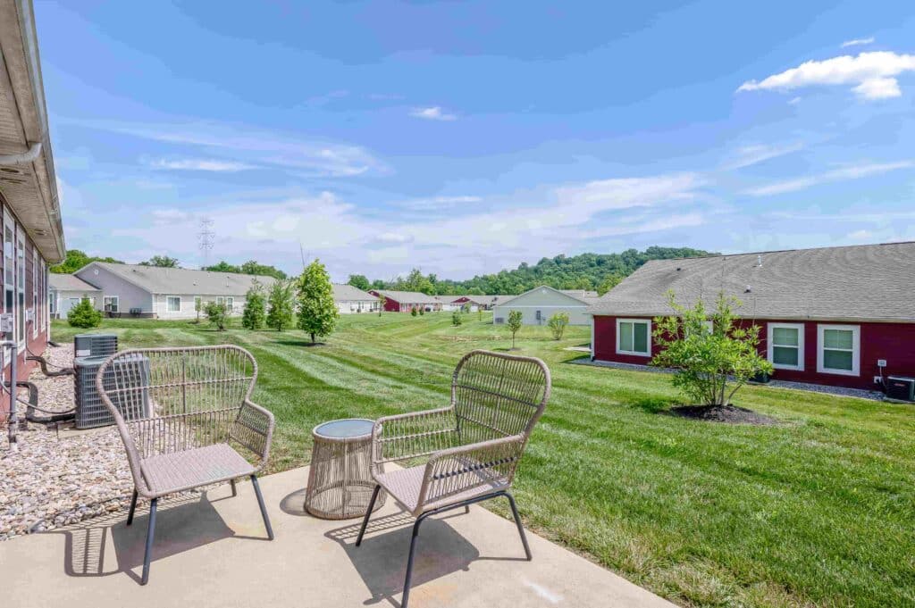 Two wicker chairs and a small table sit on a patio overlooking a grassy yard with several single-story houses under a blue sky.