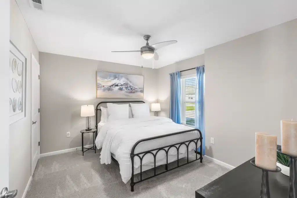 A modern bedroom with a black metal bed frame, white bedding, two side tables with lamps, a ceiling fan, a landscape painting, and a window with blue curtains.