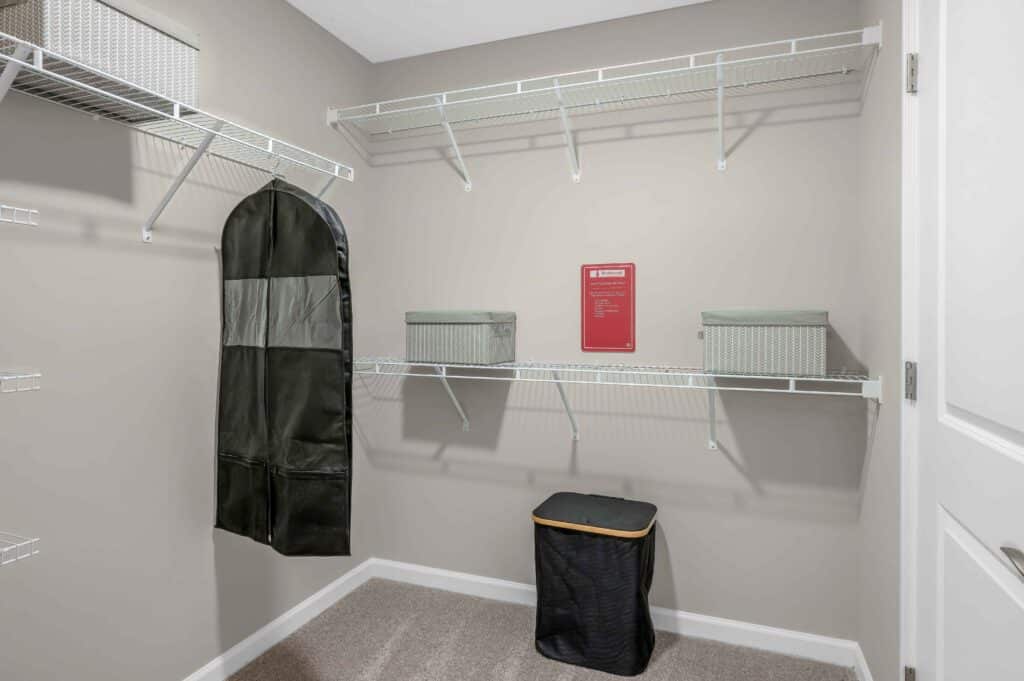 Empty walk-in closet with wire shelves, two storage bins, a black hanging garment bag, and a black laundry hamper on the carpeted floor.