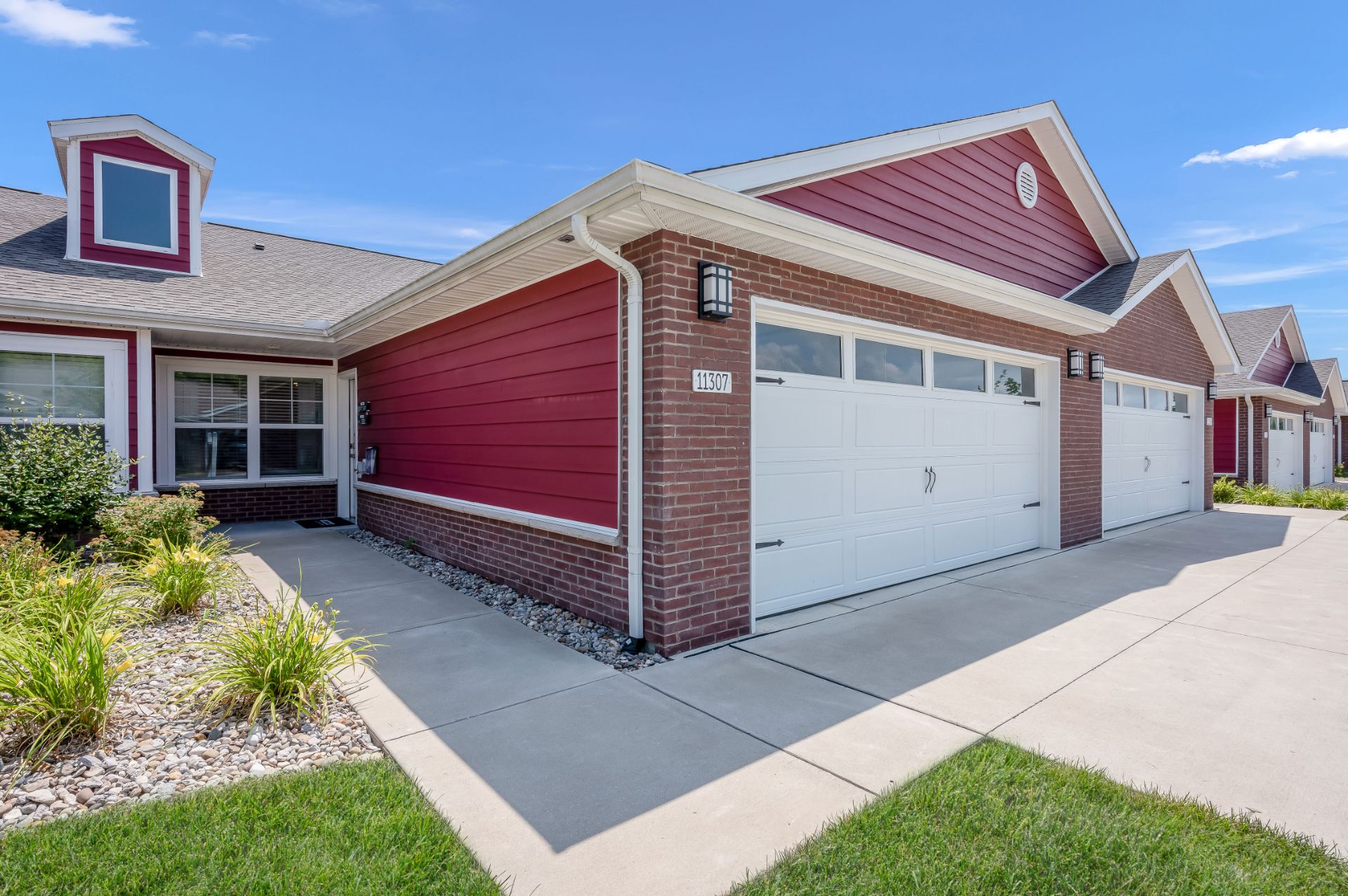Single-story red townhouse with white trim, attached two-car garage, brick accents, concrete driveway, and landscaped front yard under a blue sky.