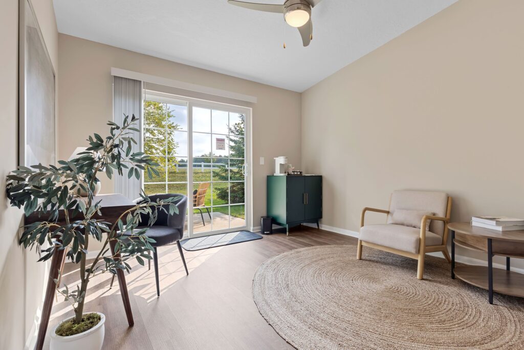 Bright room with a round rug, armchair, small table with two chairs, potted plant, and sliding glass doors leading to a patio with outdoor seating.